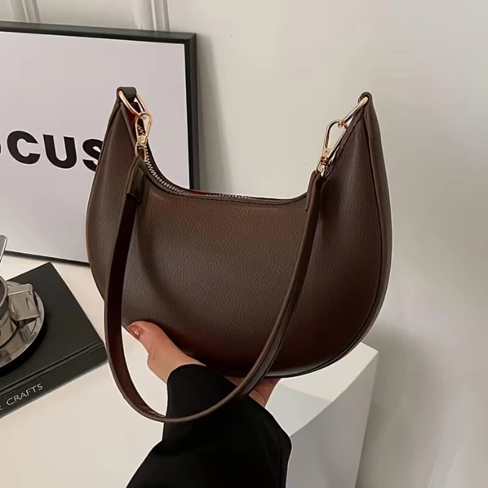 Dark brown crescent-shaped hobo shoulder bag with a minimalist design and gold hardware, held by hand against a modern white background.