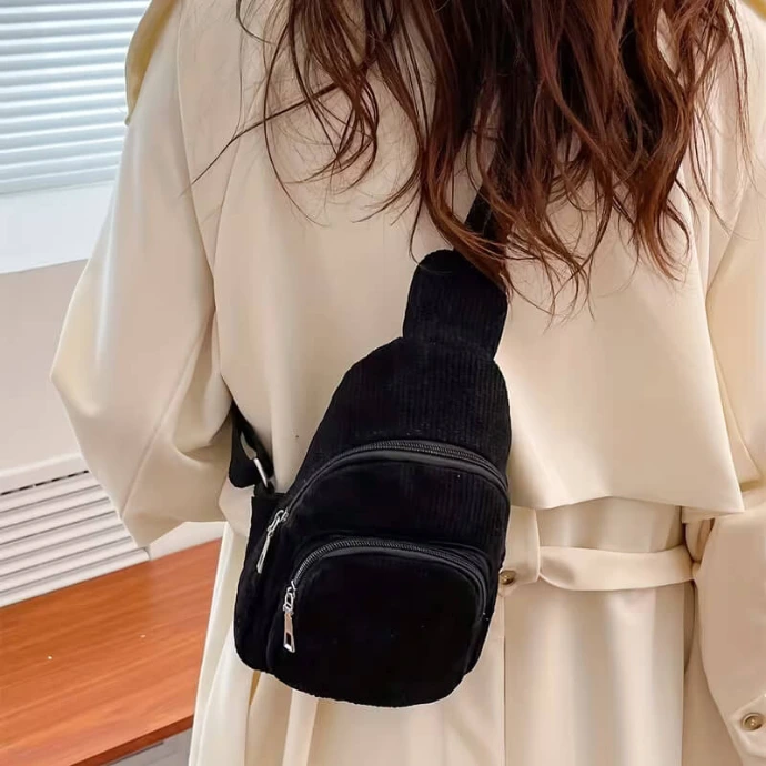 A stylish black corduroy crossbody sling bag with zippered pockets, worn by a person in a white coat.