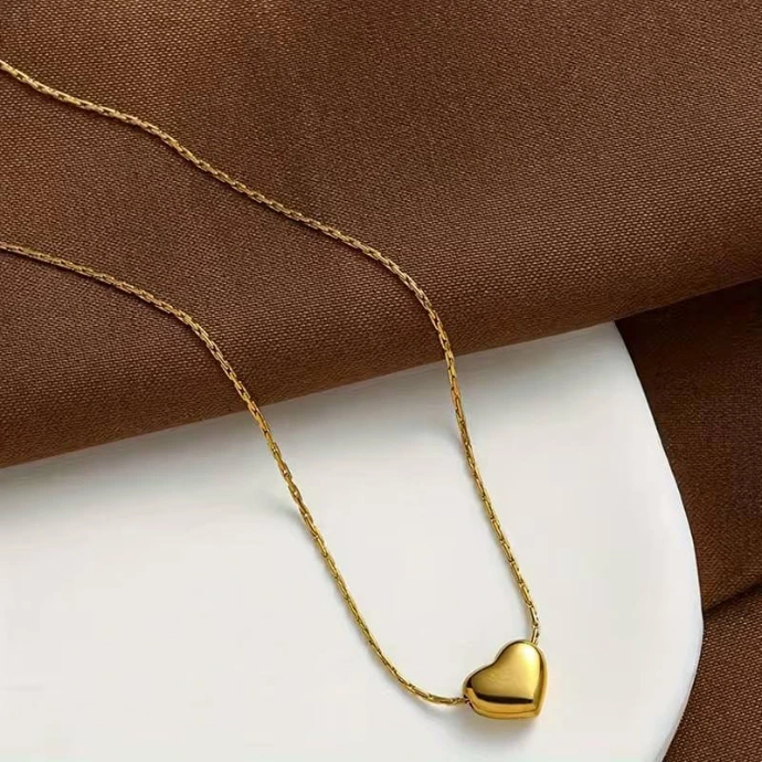 A delicate gold-tone chain necklace featuring a smooth, minimalist heart-shaped pendant, displayed on a white surface against a brown fabric background.