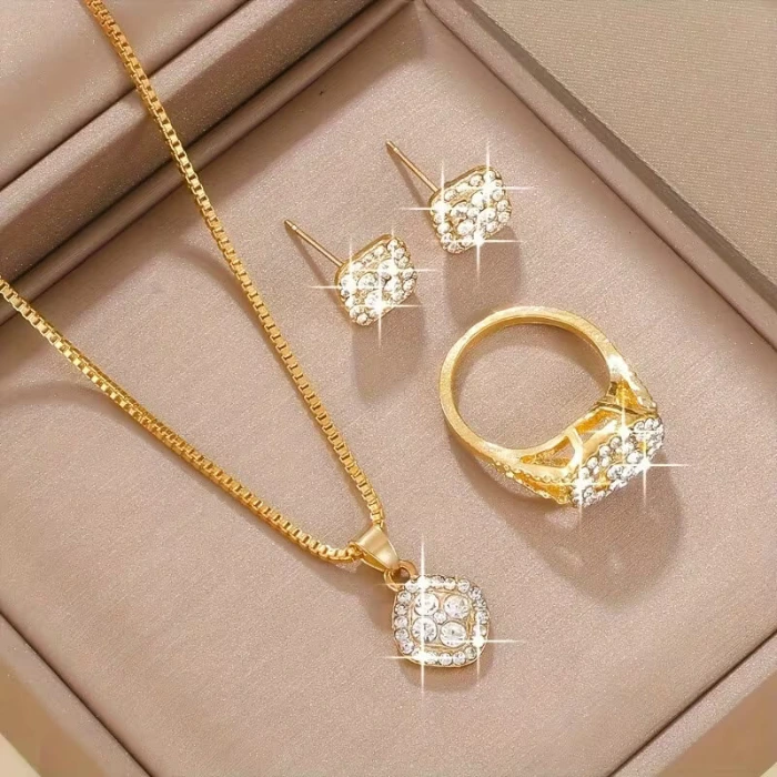 Gold jewelry set in a beige presentation box, including a crystal pendant necklace, stud earrings, and a matching ring.